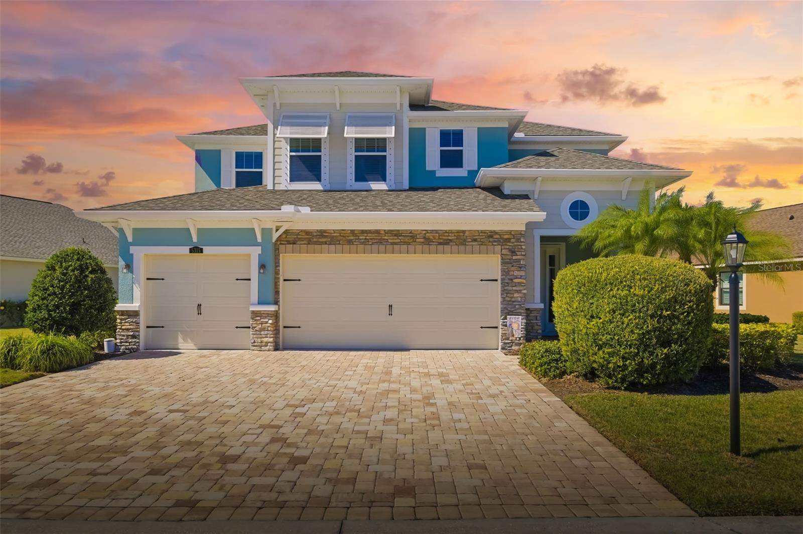Two-story blue and white house with stone accents, double garage, and broad paver driveway at sunset.