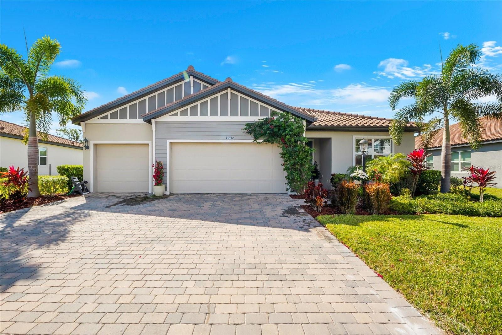 Single-story home with a tiled roof, three-car garage, paver driveway, palm trees, and landscaped front yard.