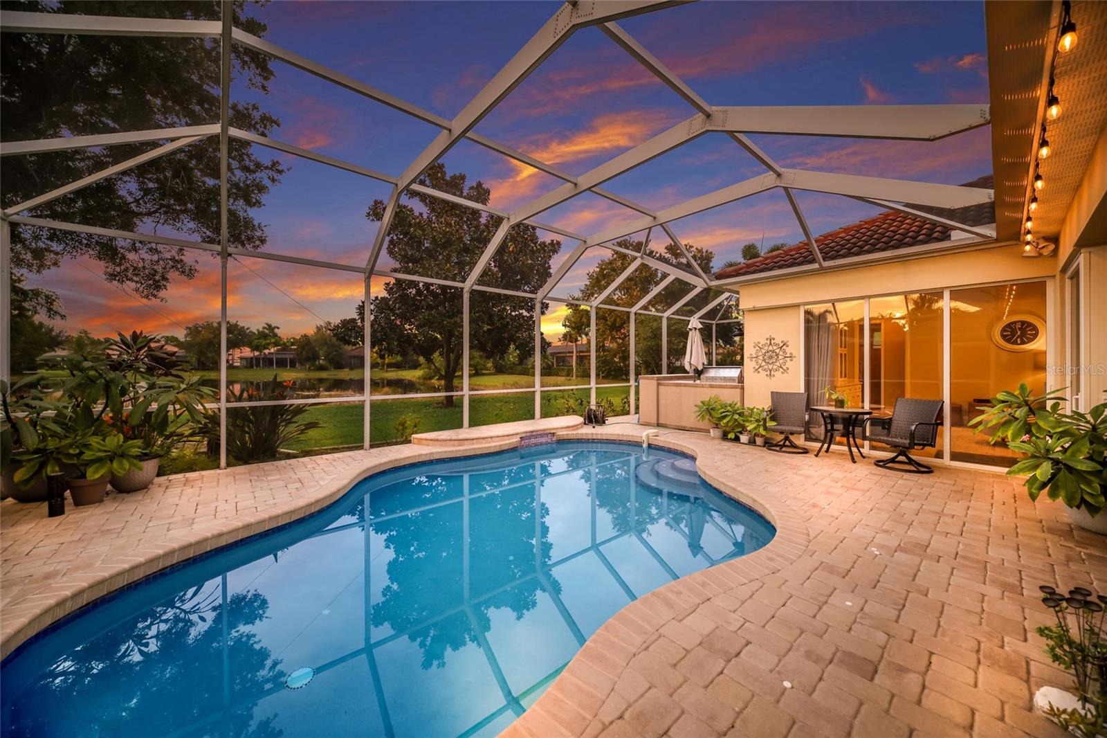 Screened backyard pool and paved patio overlooking water beneath a colorful sunset.