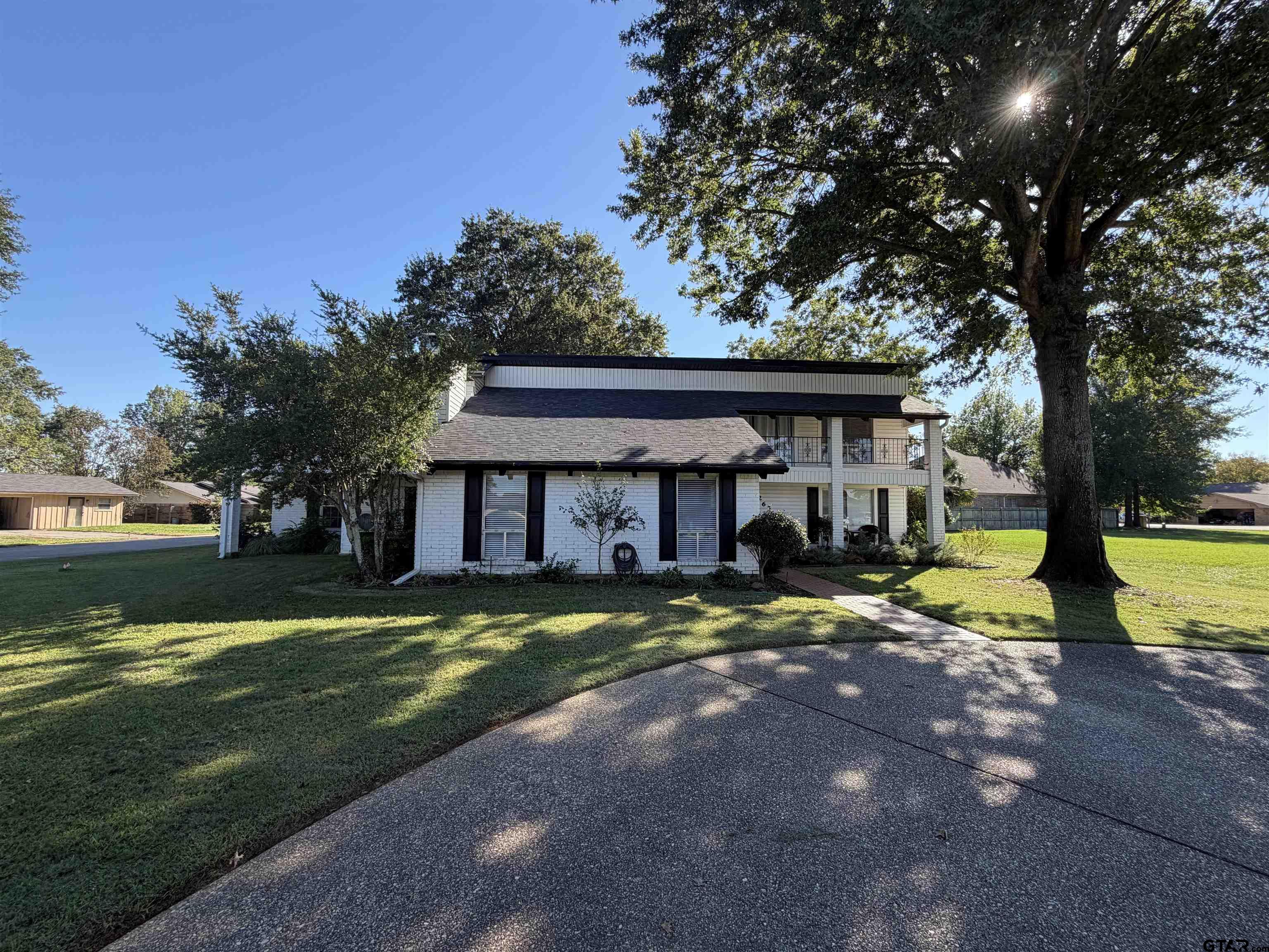 White-brick home with an upper balcony, broad lawn, and a large shade tree beside the driveway.