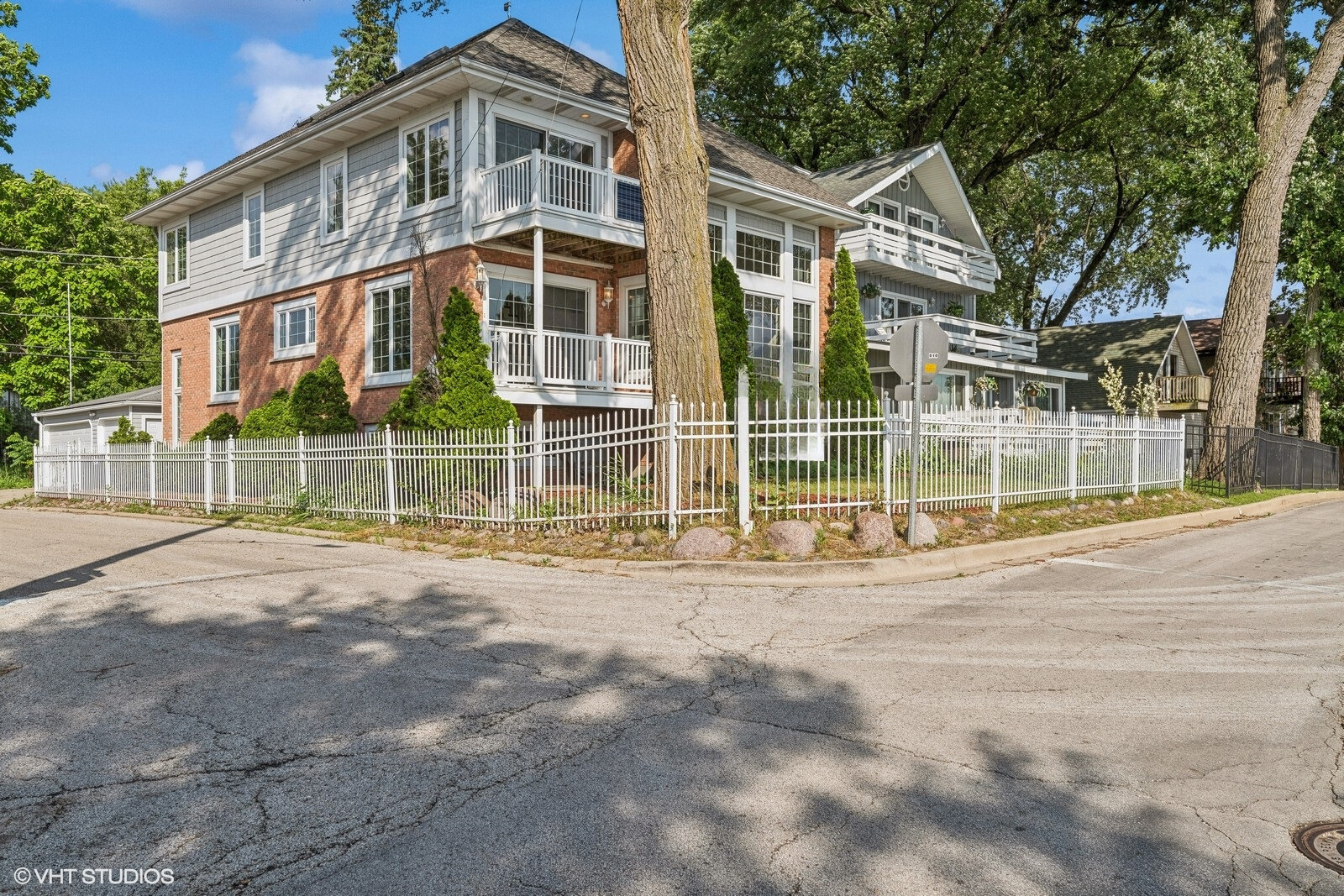 Two-story brick-and-siding home with white balconies and a picket fence beneath mature trees.