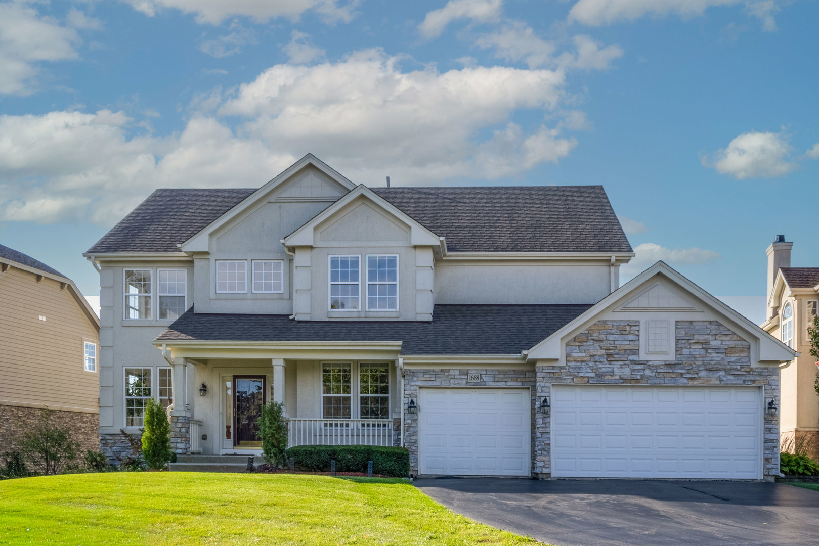 Two-story gray house with stone facade, covered porch, and two white garage doors beside a green lawn.