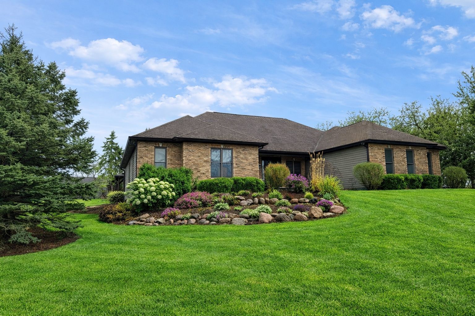 Single-story brick and gray-sided home with a rock-edged flower garden and broad green lawn.