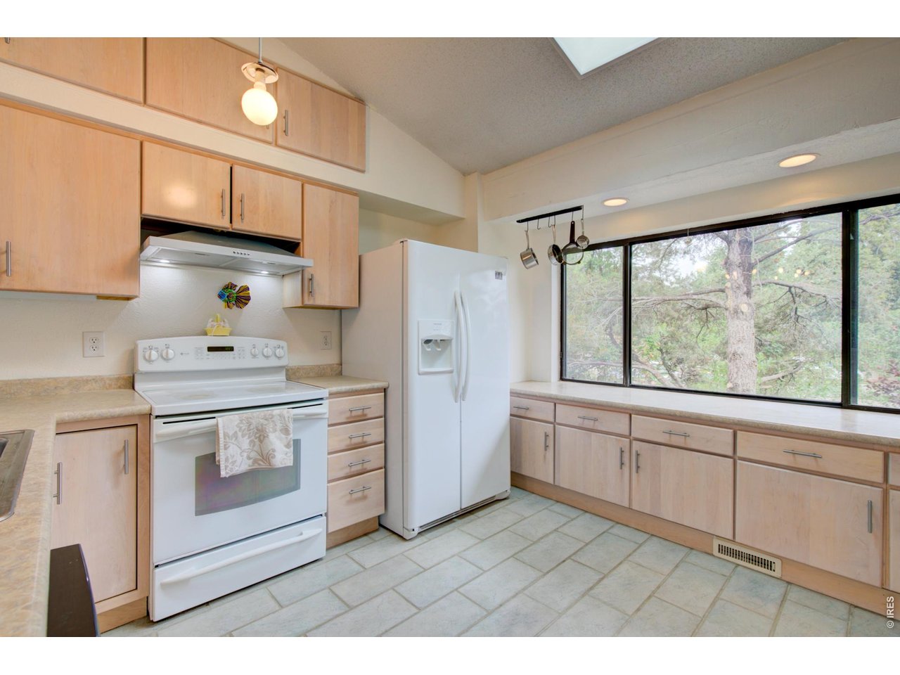 Kitchen with light wood cabinets, white appliances, and a wide window overlooking trees.