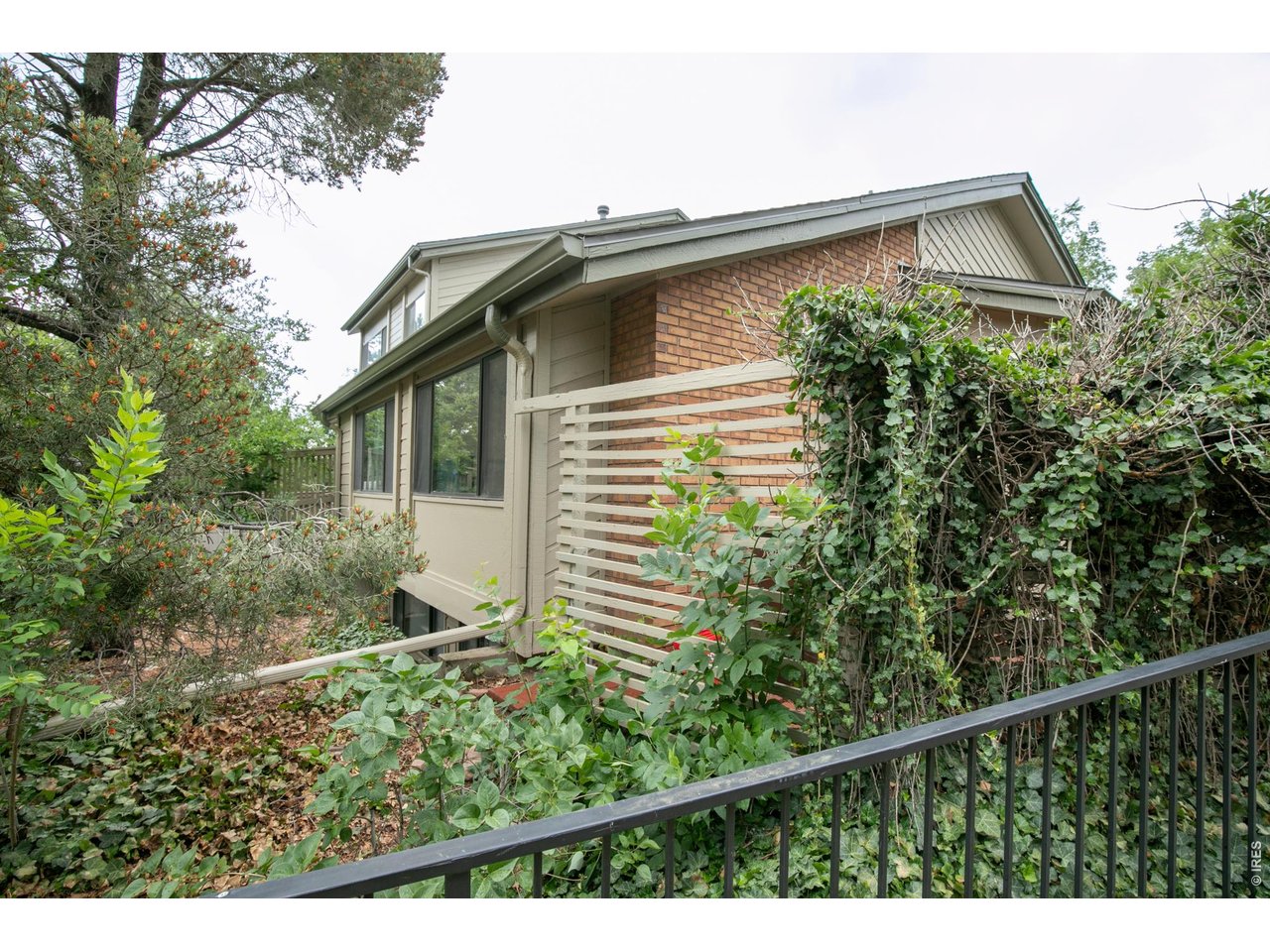 Two-story brick and beige-sided home partly screened by dense greenery, with a slatted privacy screen and railing.