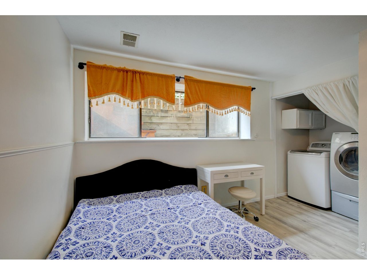 Bedroom with a blue-and-white patterned bedspread, orange window valances, and laundry appliances in an alcove.