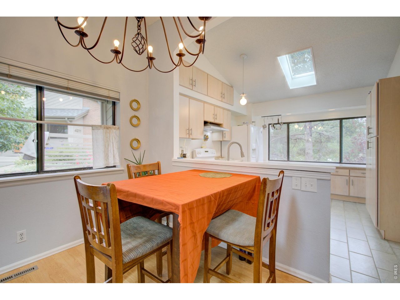 Dining area with an orange-covered table, chandelier, large windows, and skylight beside the kitchen.