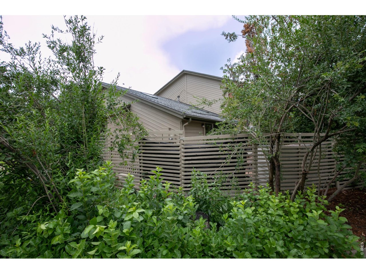 Beige-sided home behind a slatted privacy fence, surrounded by dense greenery.