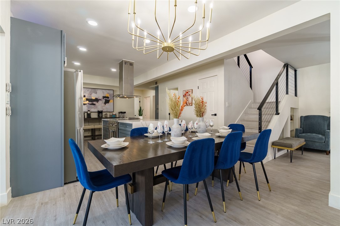 Dining room with a dark set table, blue chairs, gold chandelier, and open kitchen beside a staircase.