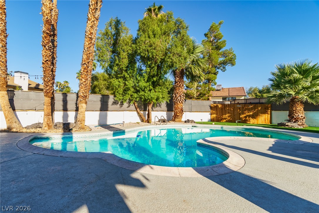 Backyard pool with a curved turquoise swimming area, concrete deck, palm trees, and wooden privacy screen.