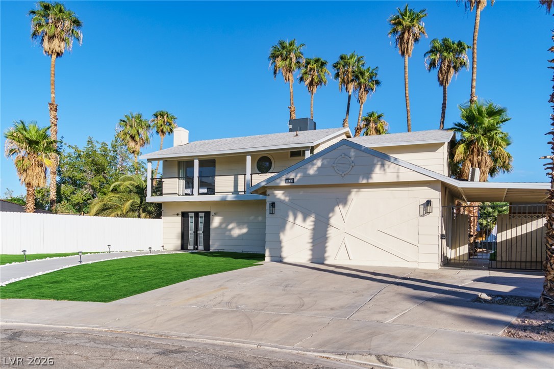 Two-story light-colored house with a front garage, covered carport, broad driveway, and palm trees.