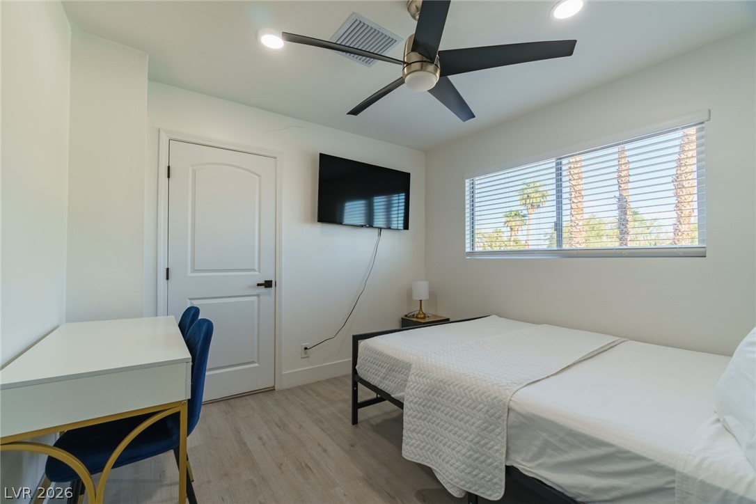 Furnished bedroom with a white bed, desk, blue chair, and window overlooking palm trees.