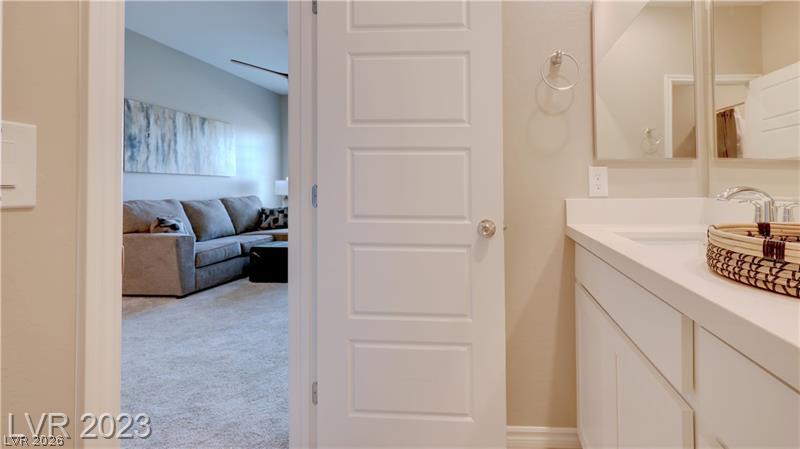 Bathroom vanity beside an open door looking into a carpeted room with a gray sectional sofa.