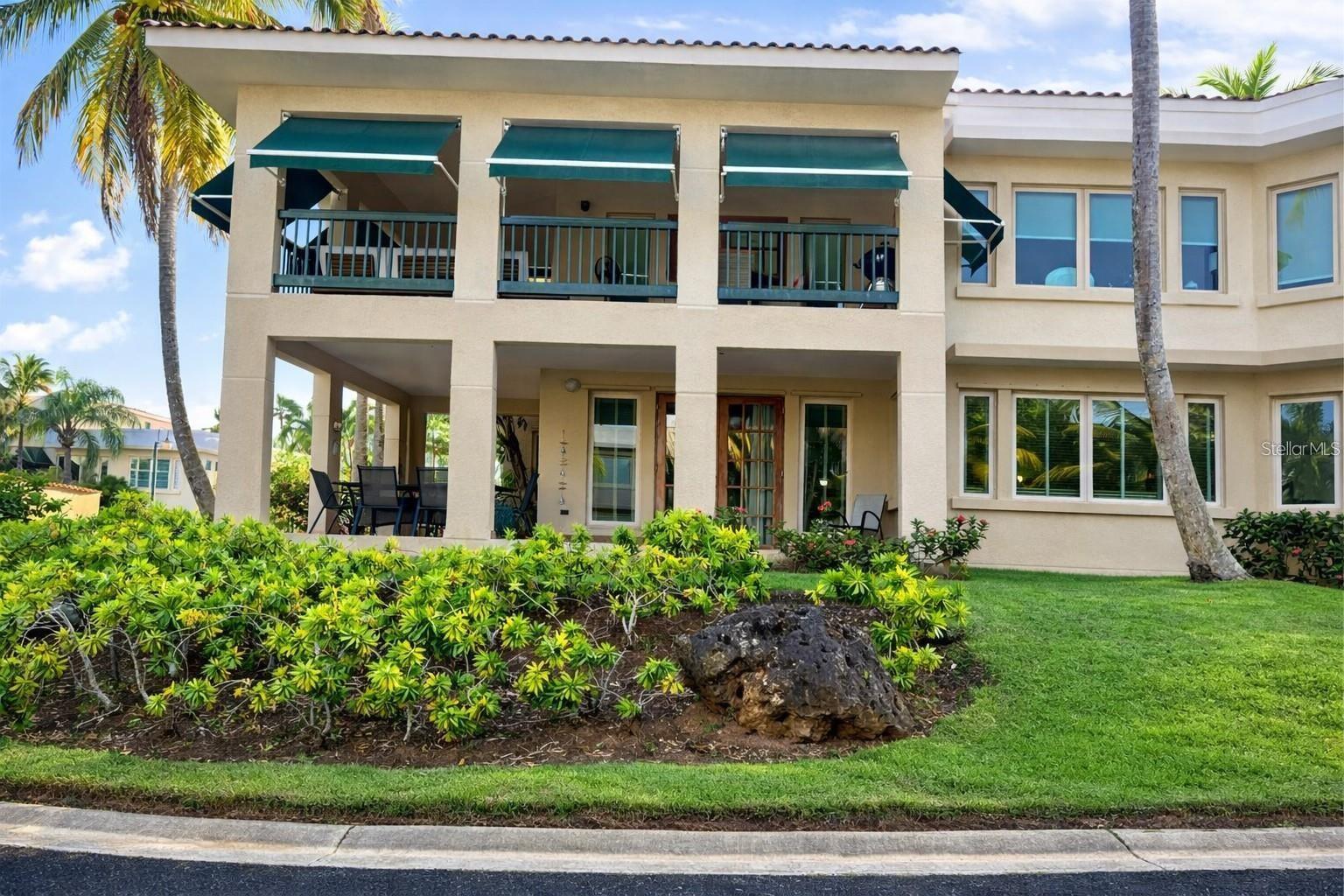 Two-story beige residence with teal balcony awnings, a covered patio, tropical landscaping, and palm trees.