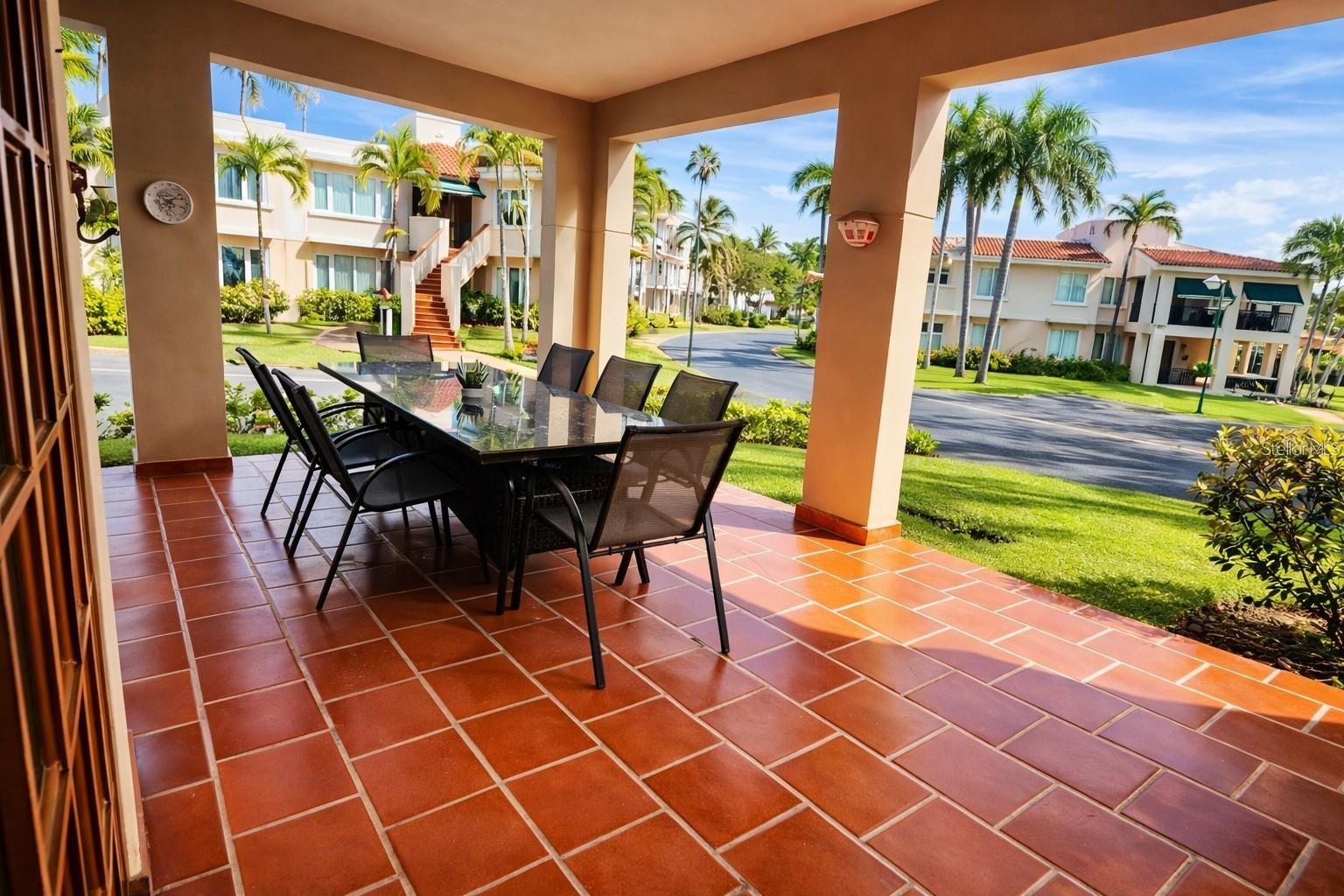 Covered tiled patio with a glass-top dining table overlooking palm-lined villas.