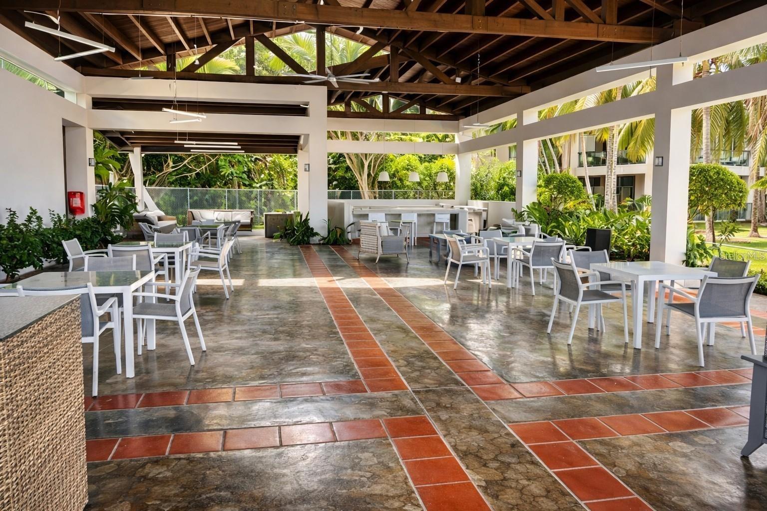 Open-sided community pavilion with white dining tables, exposed wooden roof trusses, and tropical landscaping.