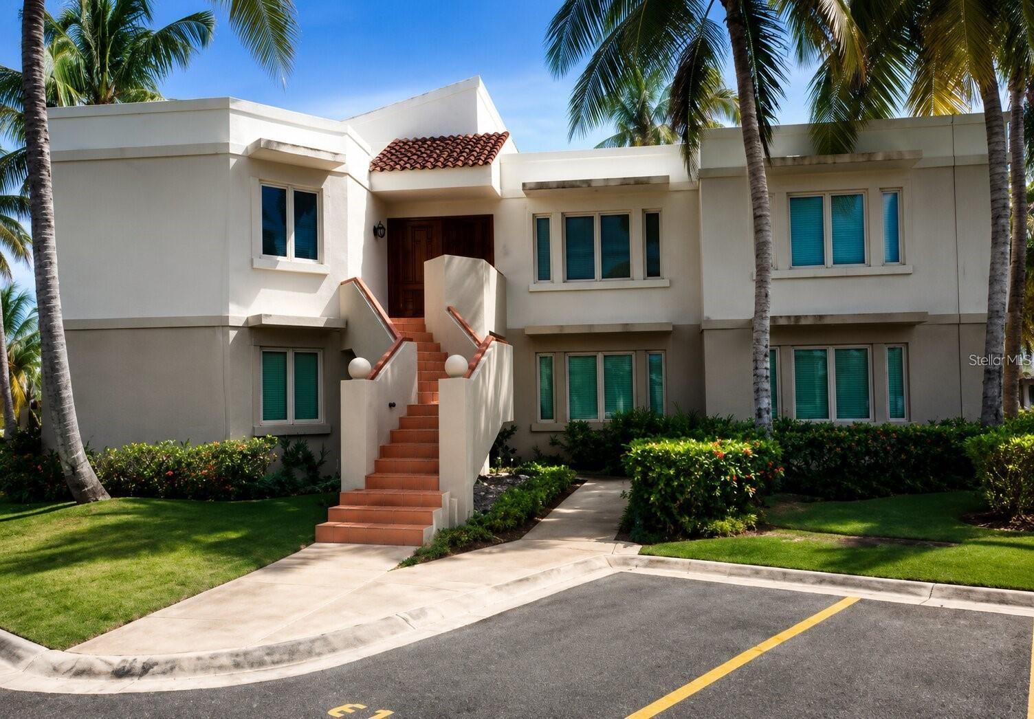 White two-story condominium building with a terracotta-tiled entry and staircase, framed by palm trees.