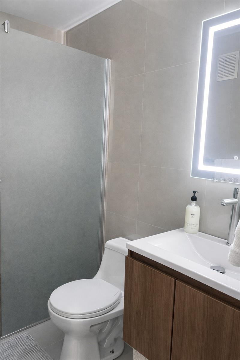 Bathroom with a glass shower, white toilet, wood vanity, and illuminated mirror.