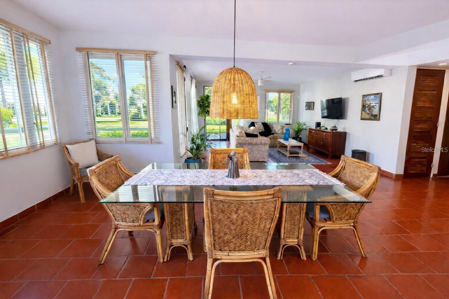 Dining and living room with wicker furniture, terracotta tile floors, and large windows overlooking greenery.