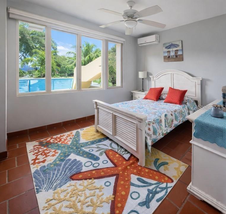 Bedroom with a louvered white bed and colorful sea-life rug beside windows overlooking a pool.