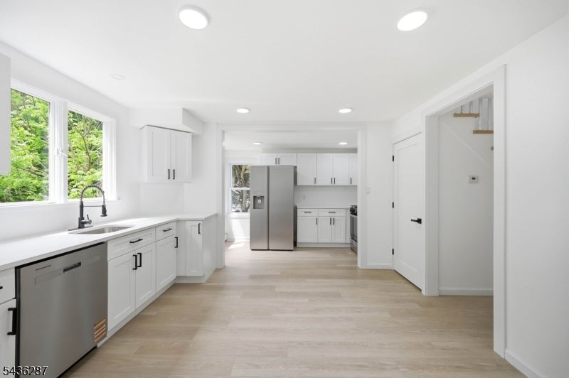Bright white kitchen with pale wood floors, stainless steel appliances, and windows overlooking greenery.