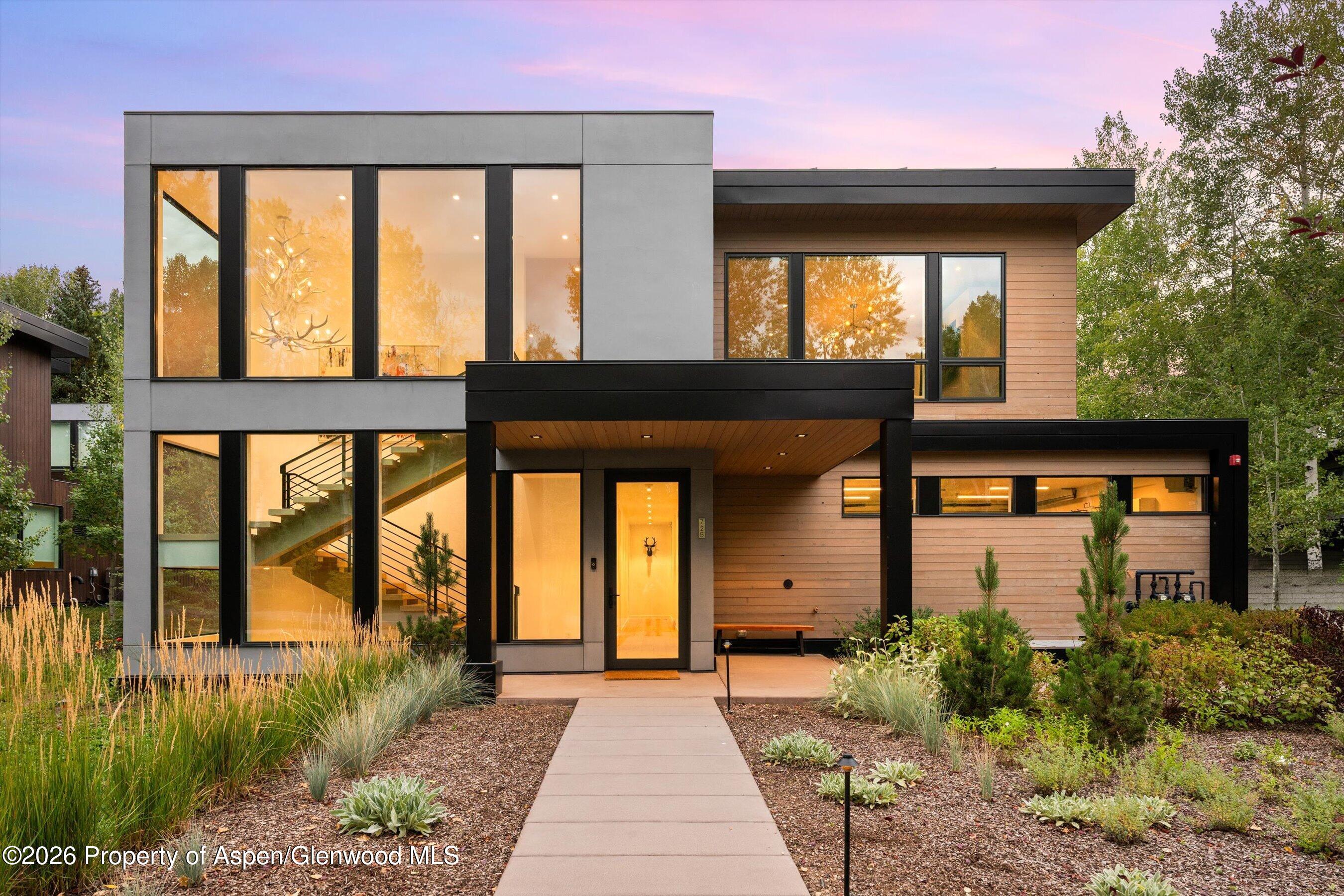 Modern two-story home with large windows, wood siding, and a landscaped front walkway at dusk.