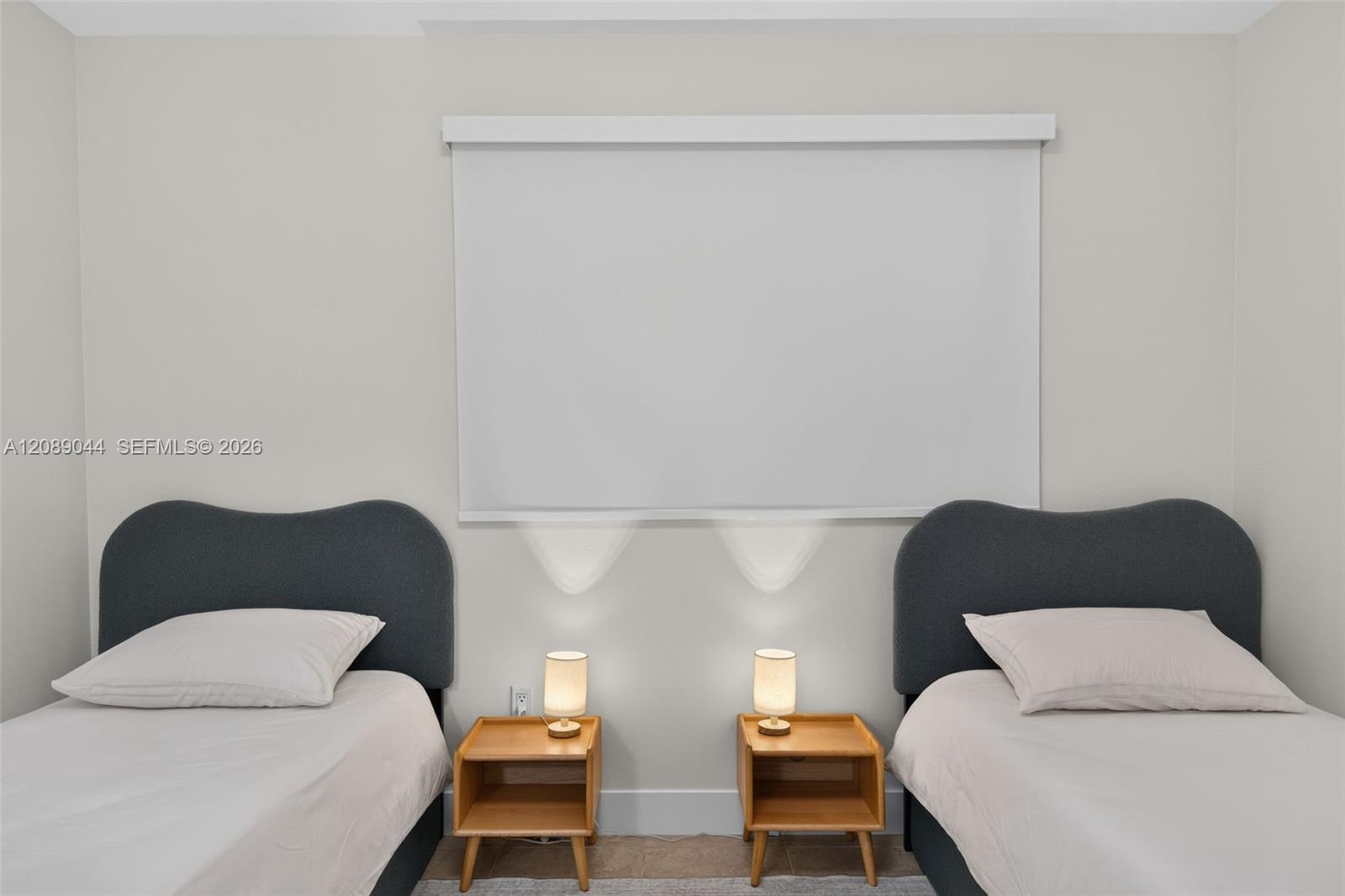 Bedroom with two twin beds, matching wooden nightstands, and a closed roller shade.