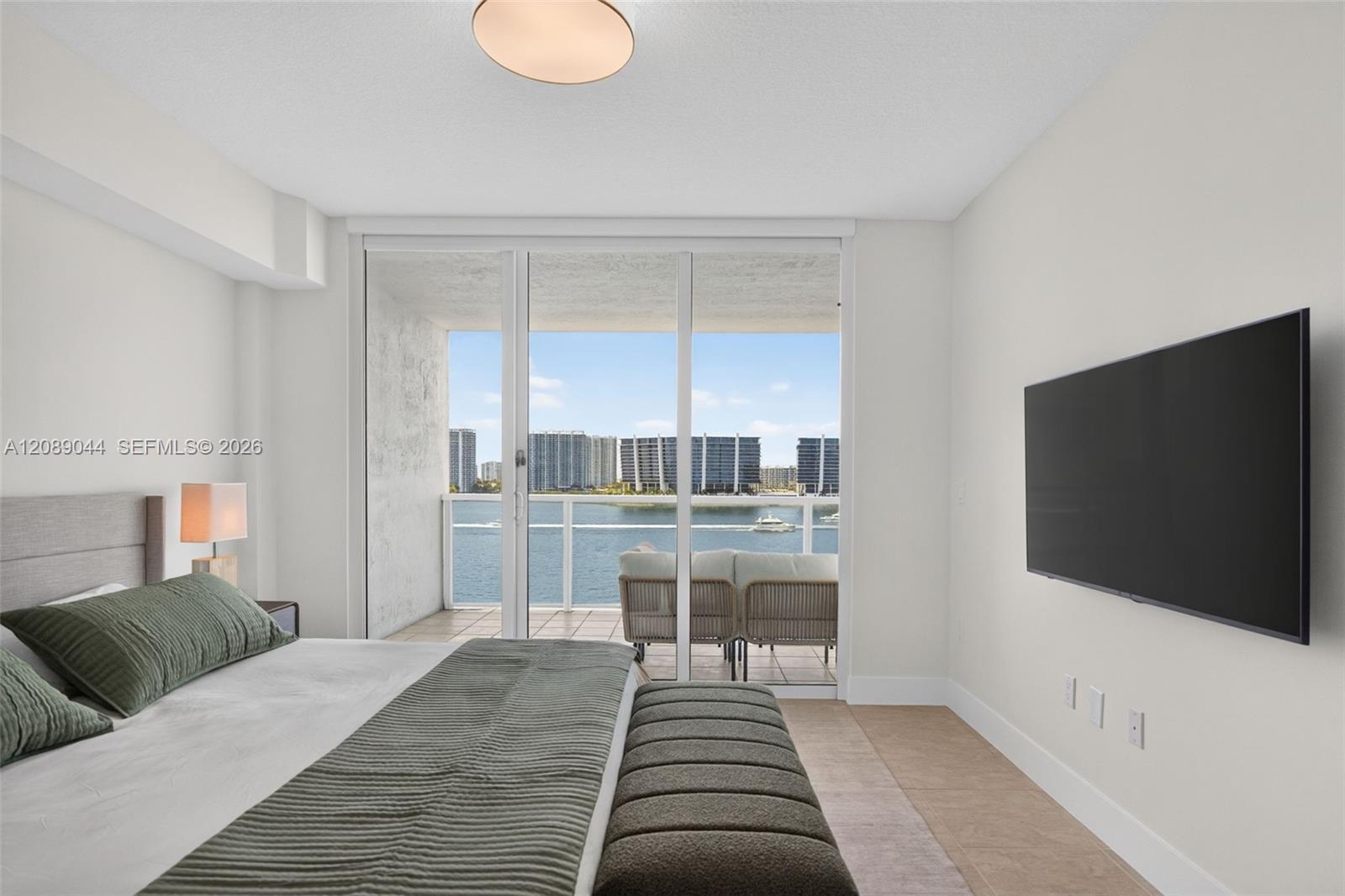 Bedroom with balcony doors overlooking water and a city skyline.