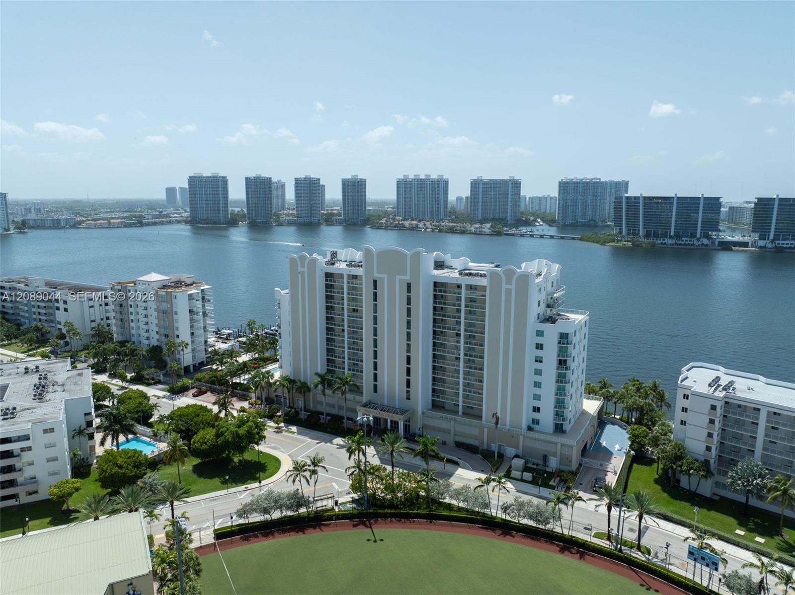 Aerial waterfront view with a white condominium, bay, and distant high-rise skyline.