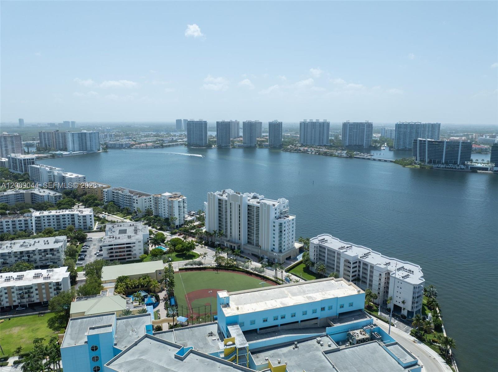 Aerial view across a bay toward waterfront towers, with a sports field and buildings in the foreground.