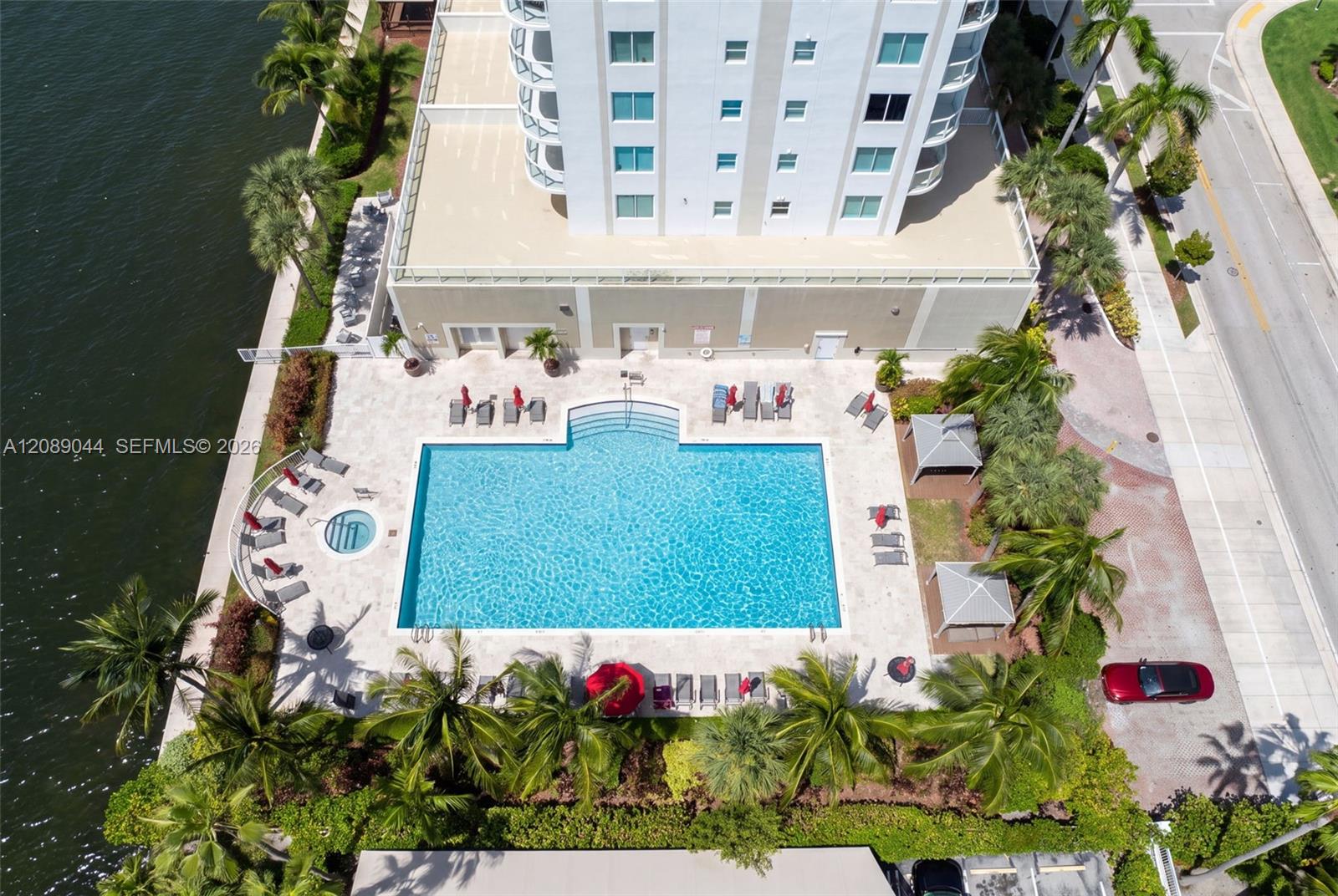 Aerial view of a condominium pool and spa beside a waterway, surrounded by palms and lounge chairs.