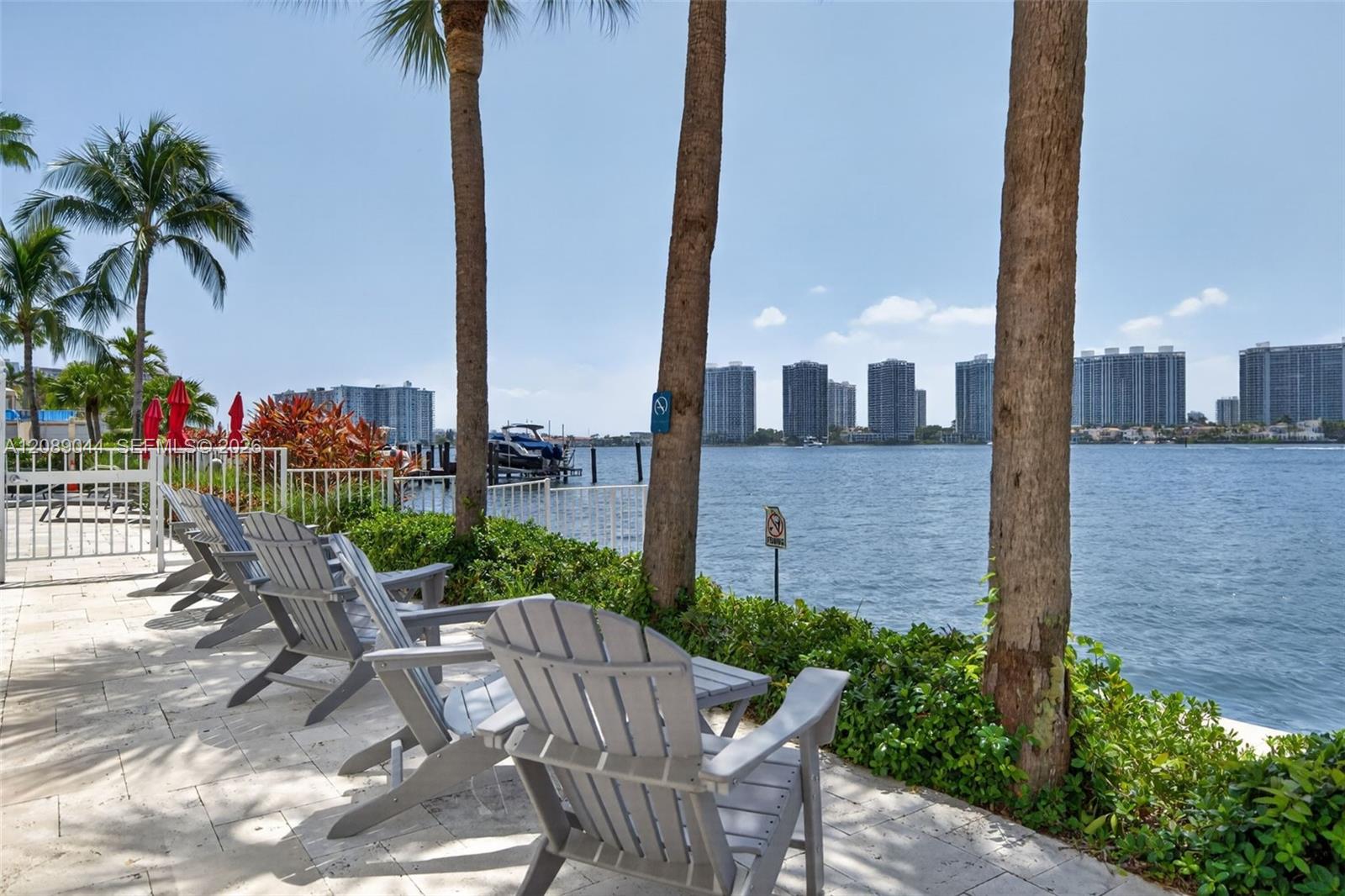Waterfront patio with Adirondack chairs, palm trees, and a distant city skyline across the water.