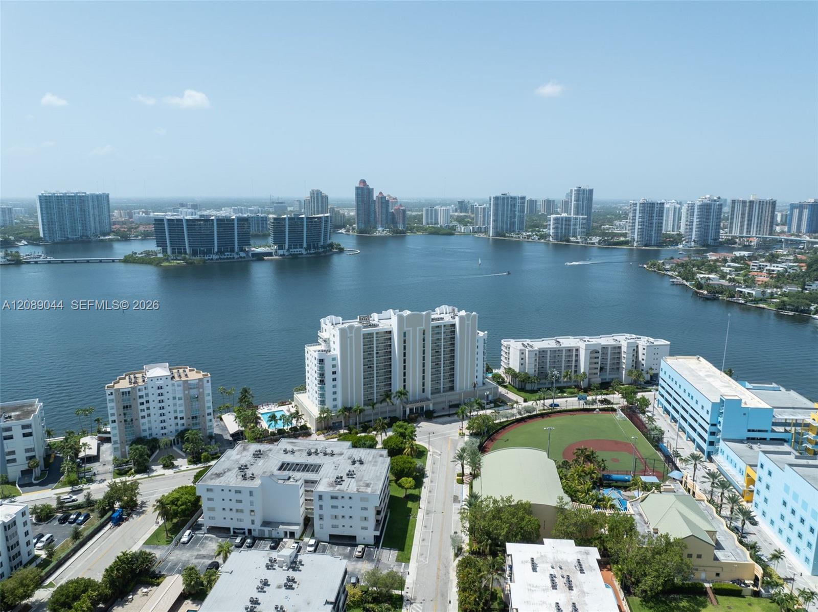 Aerial view of waterfront buildings along a bay, with a baseball field and distant skyline.