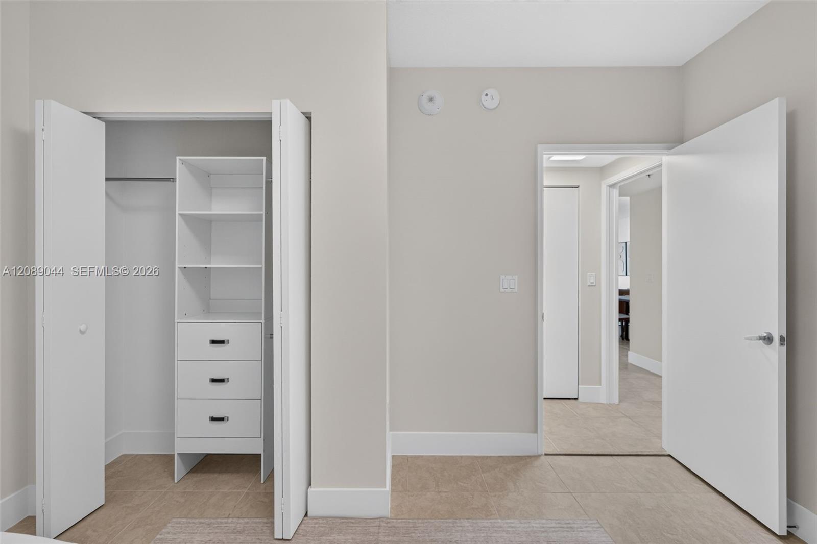 Empty room with an open built-in closet featuring white shelves and drawers, beside a tiled hallway.