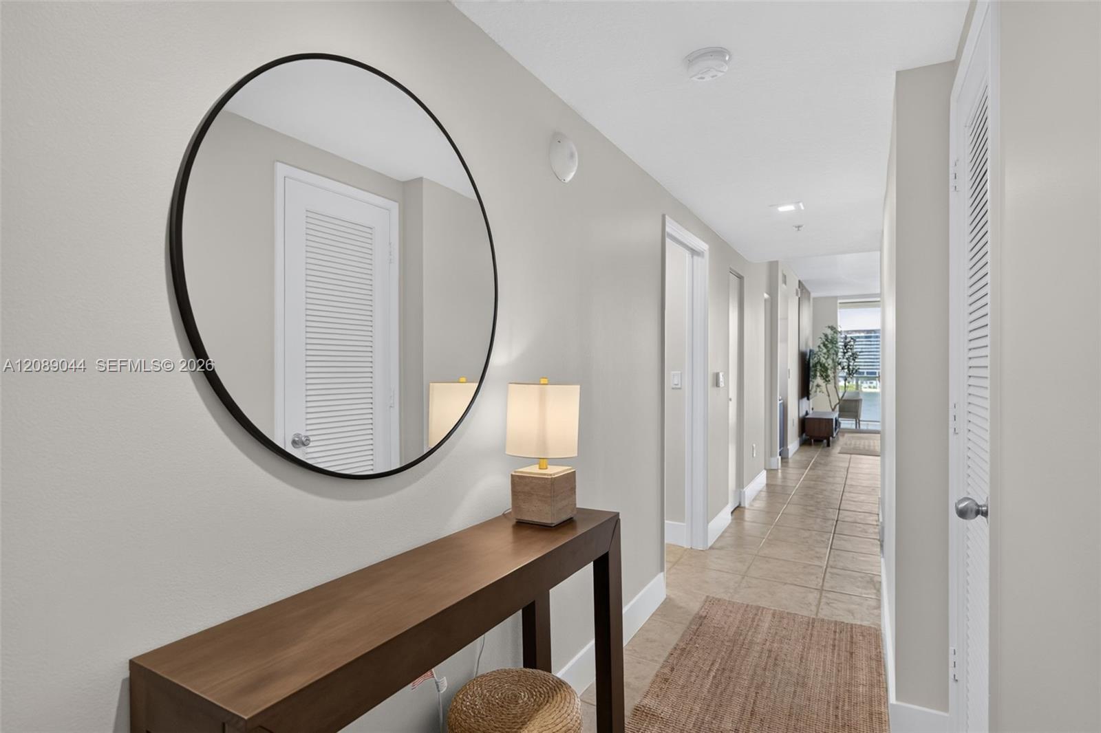 Furnished apartment hallway with a round mirror, wooden console table, and tiled floor.