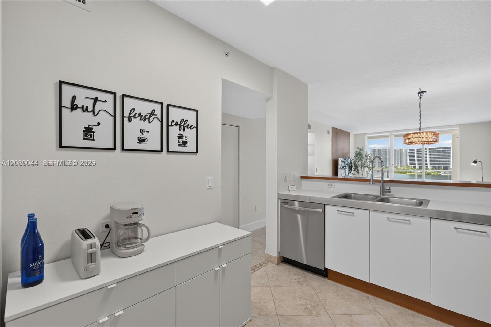 Modern white kitchen with a double sink, stainless-steel dishwasher, and views of water and high-rises.