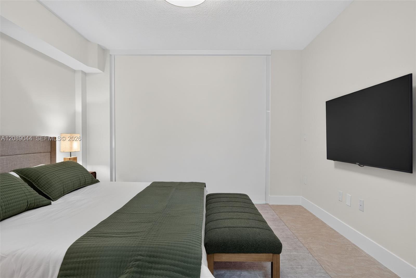 Bedroom with dark green bedding, a padded bench, and a wall-mounted television.