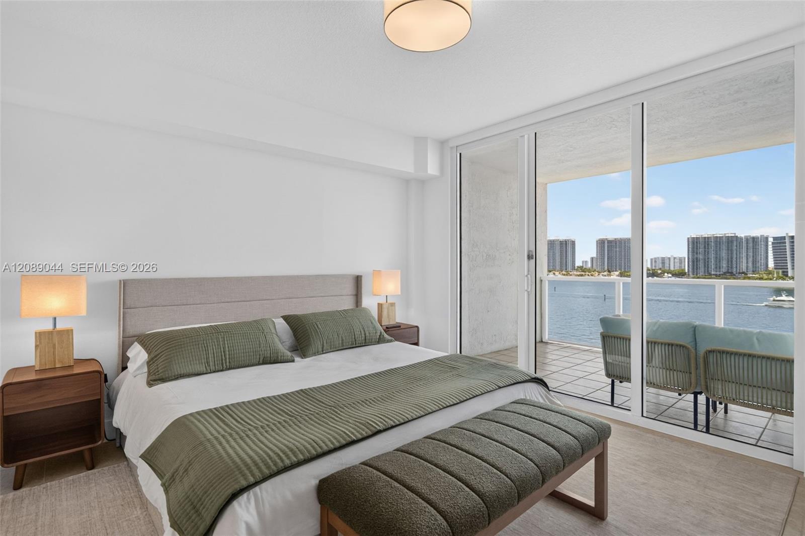 Staged bedroom with a balcony overlooking the water and high-rise buildings.
