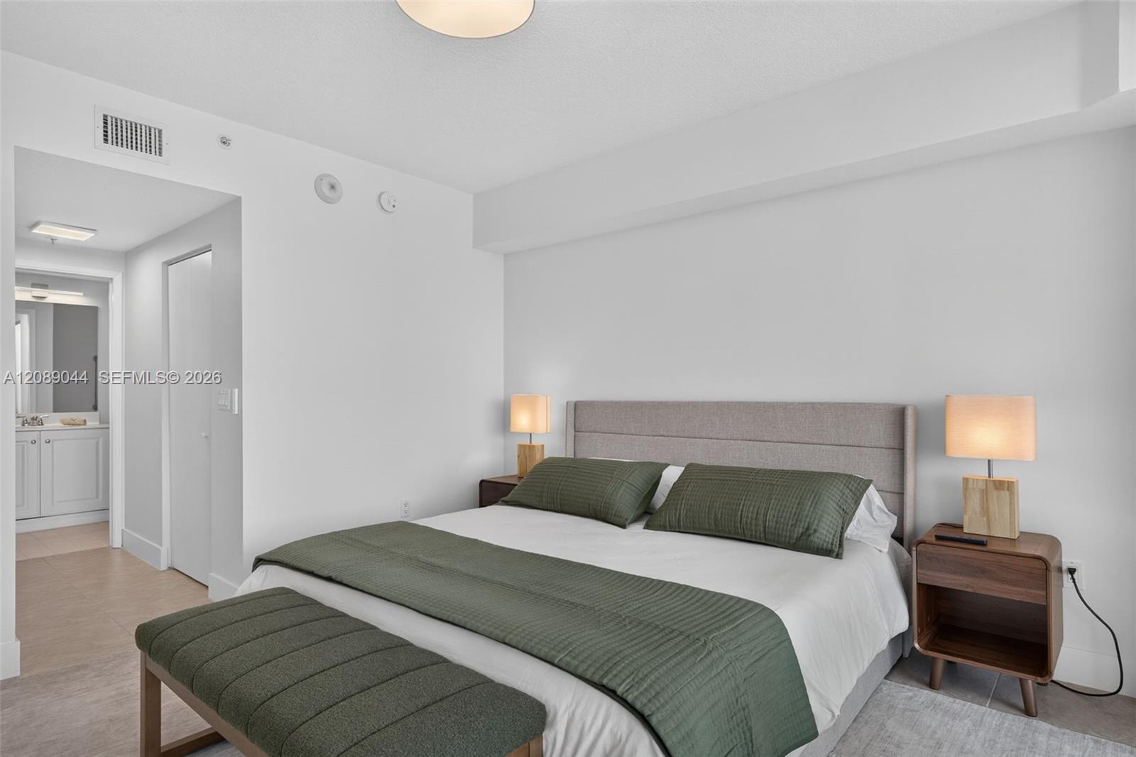 Staged bedroom with a king bed, green bedding, matching bedside lamps, and an ensuite vanity.