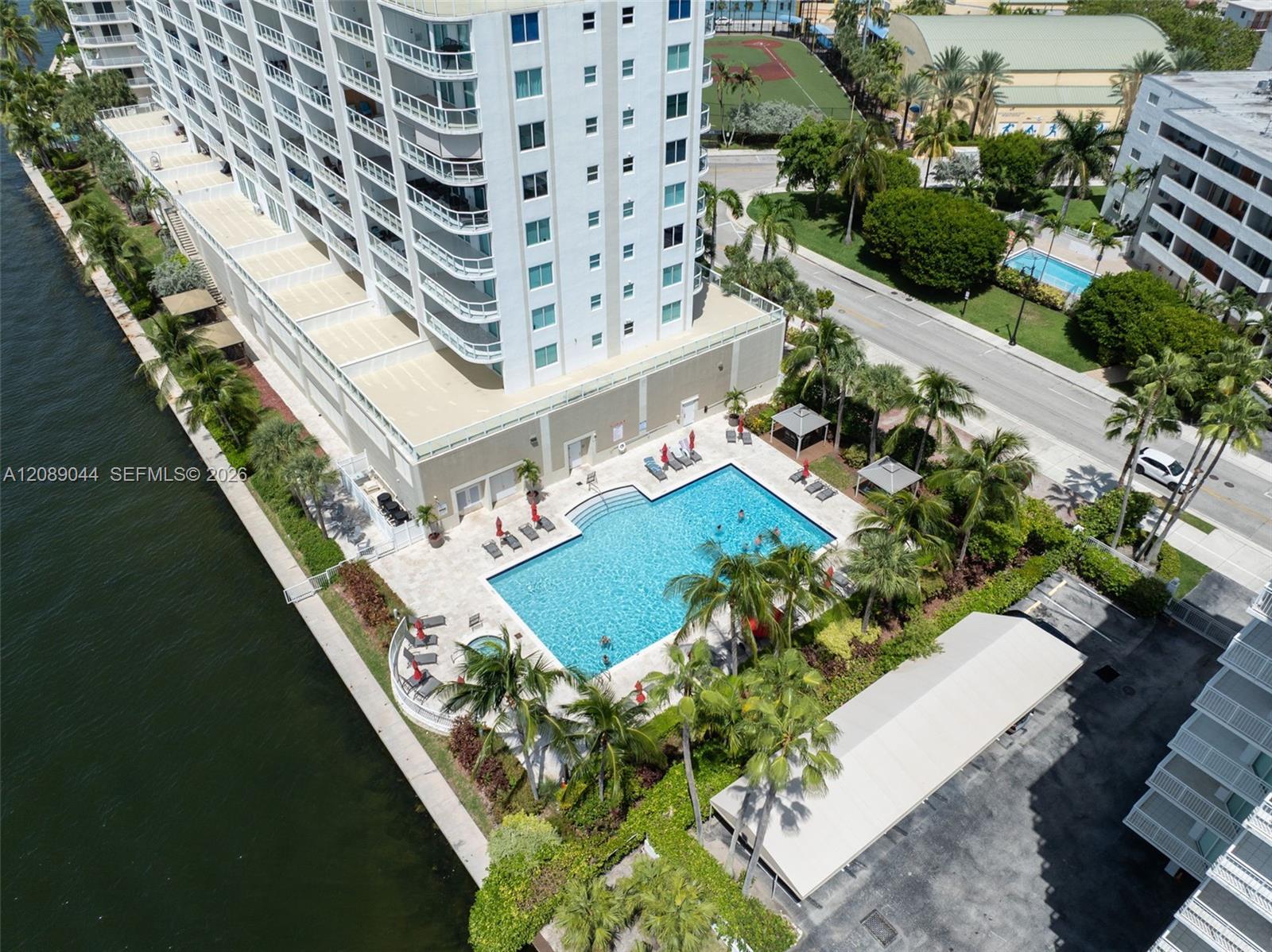 Aerial view of a condominium beside a canal, with a palm-lined swimming pool and tennis court.