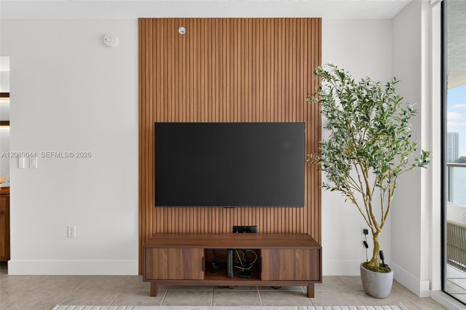Living room with a television on a wood-slat wall and a potted tree beside balcony doors.
