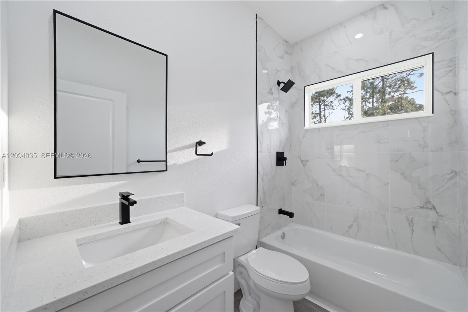 Modern bathroom with marble-look tub surround, black fixtures, and a window overlooking trees.