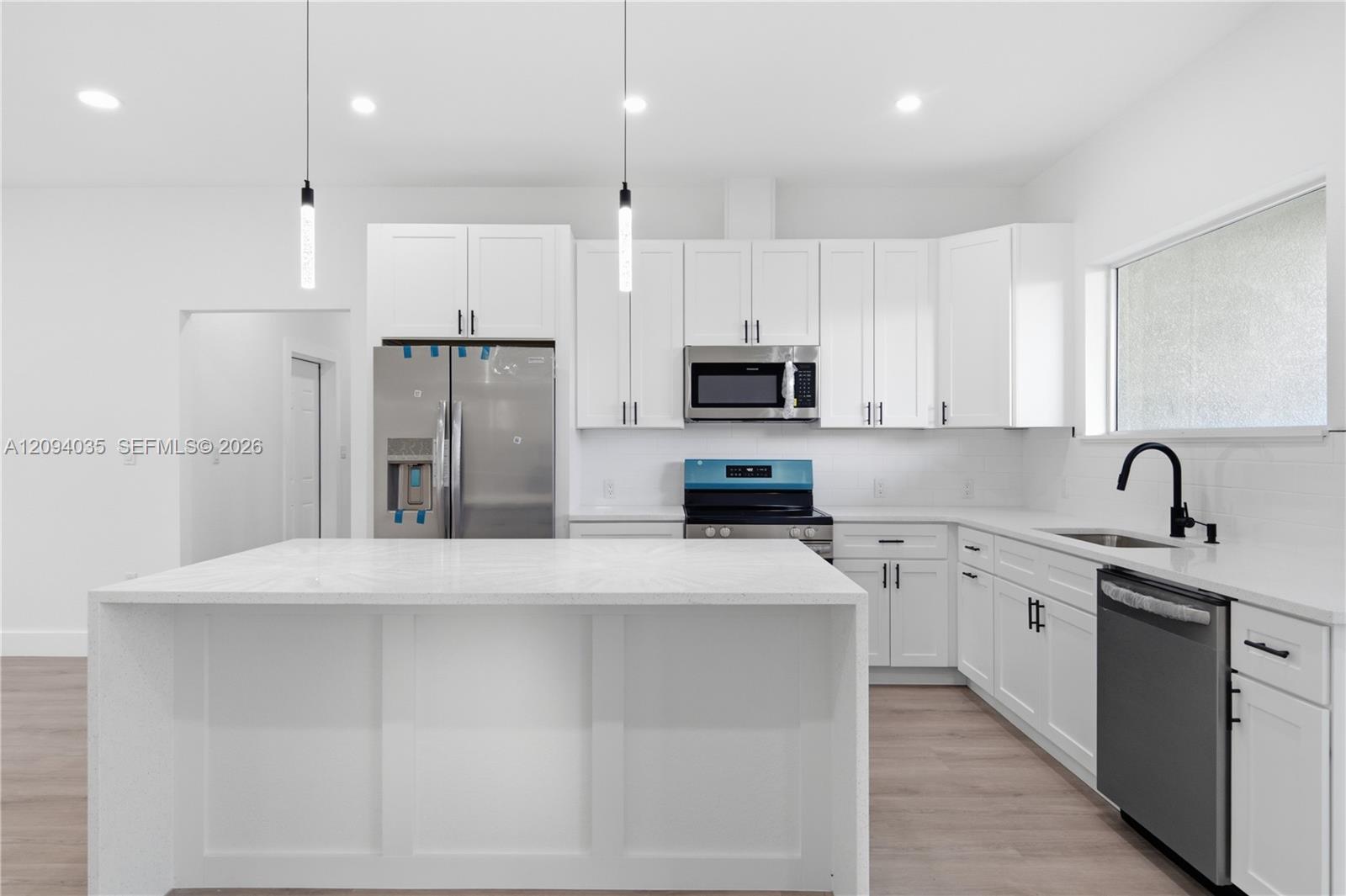 Modern white kitchen with a large island, stainless steel appliances, and black faucet beneath a wide window.