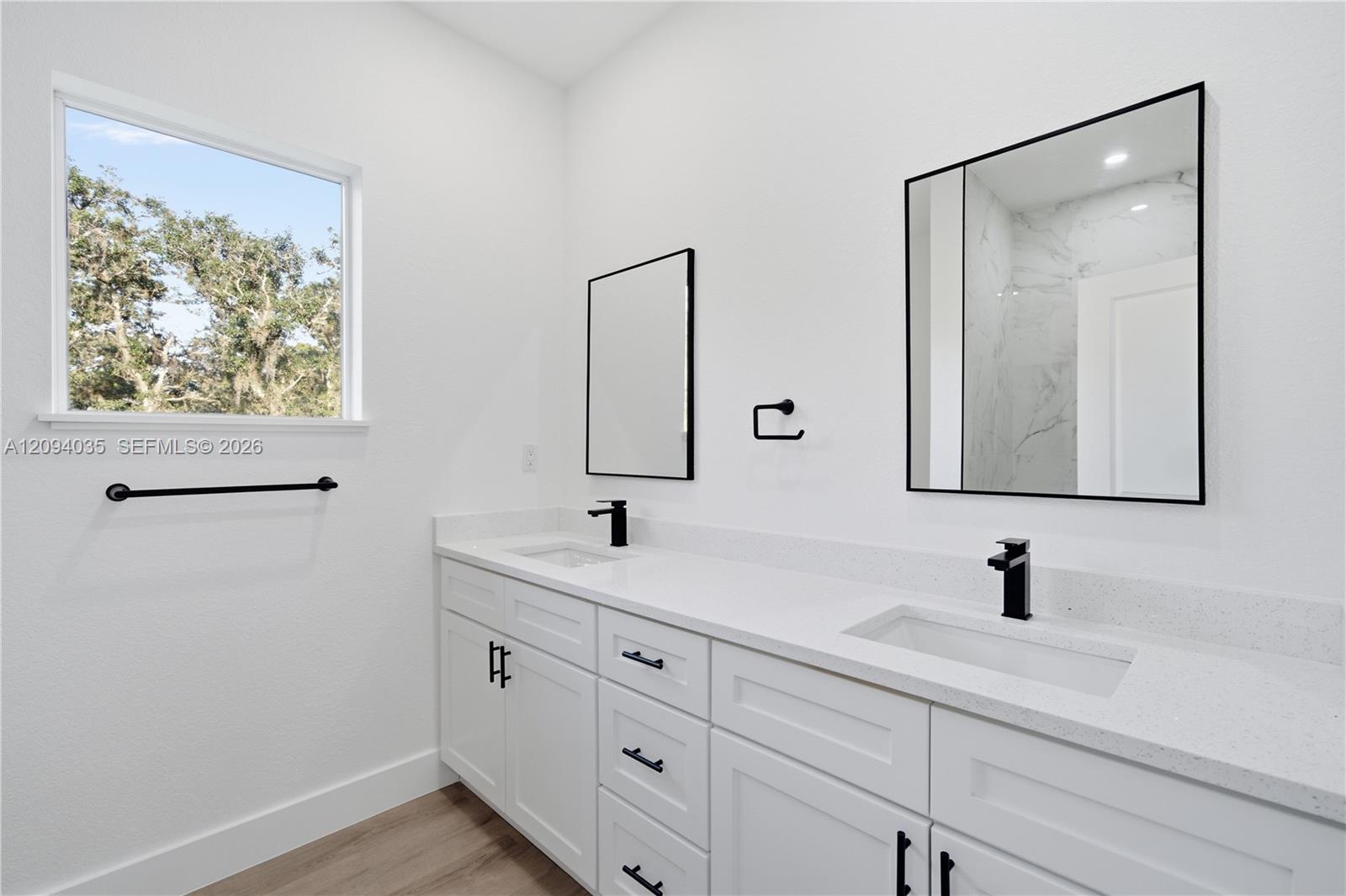 Modern bathroom with a white double-sink vanity, black fixtures, two mirrors, and a window overlooking trees.