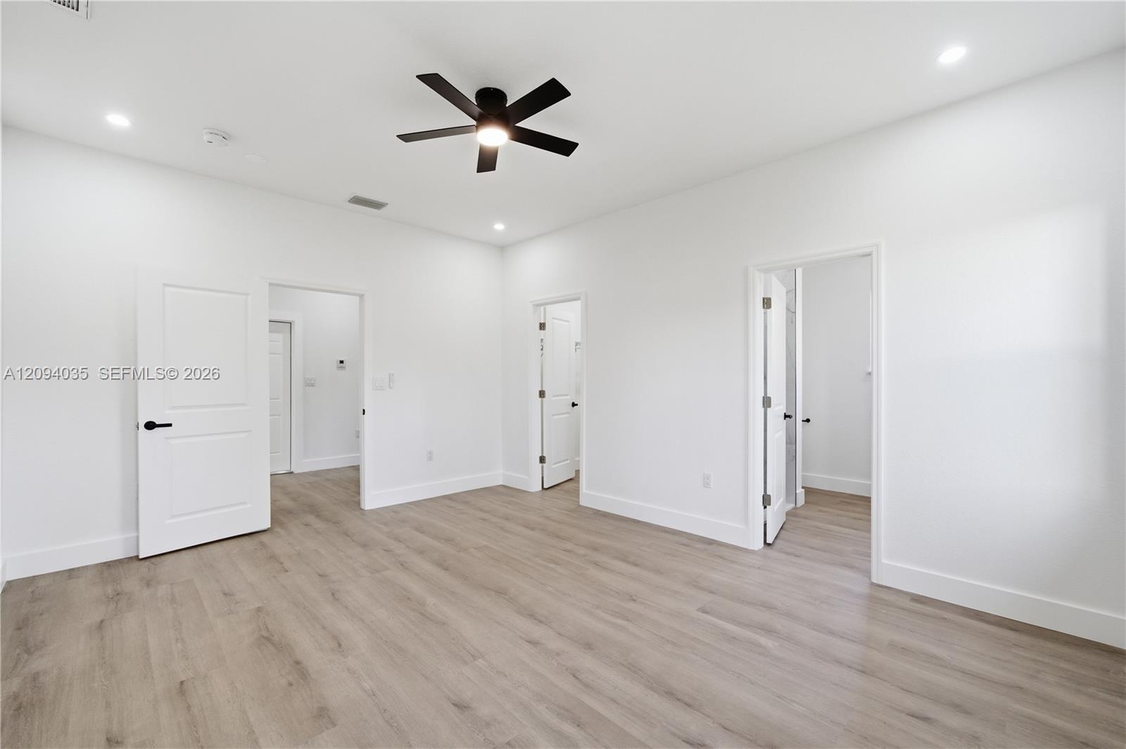 Empty bright room with light wood-look flooring, multiple doorways, and a dark ceiling fan.