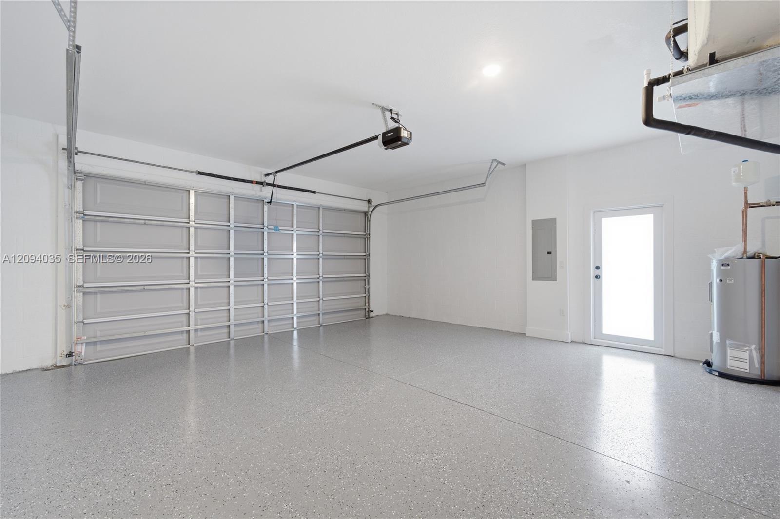 Empty garage with a large overhead door, speckled gray floor, side entry door, and water heater.