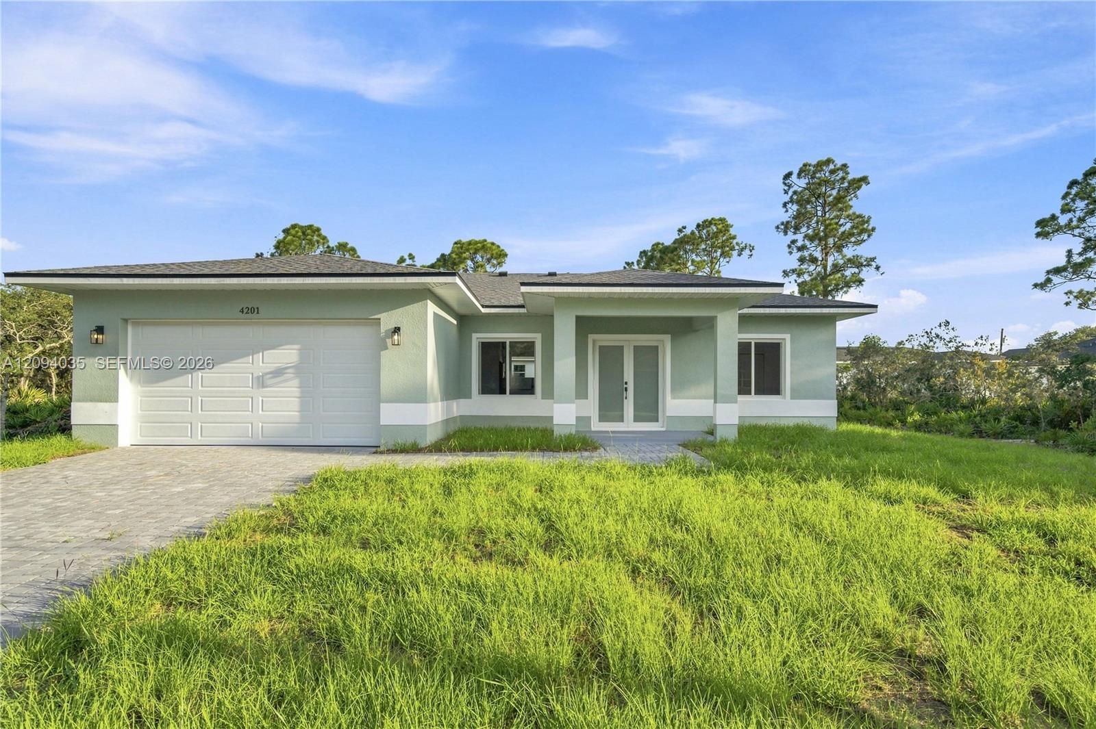 Single-story pale green house with a white double garage door, covered entry, and paver driveway.
