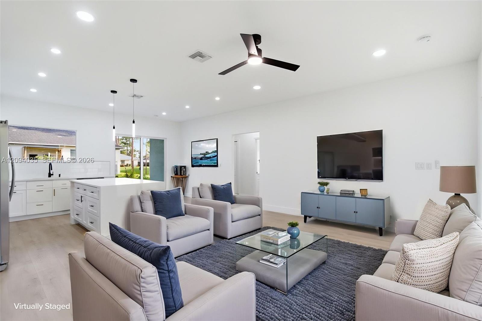 Virtually staged living room with white sofas, gray rug, glass coffee table, and open kitchen island.
