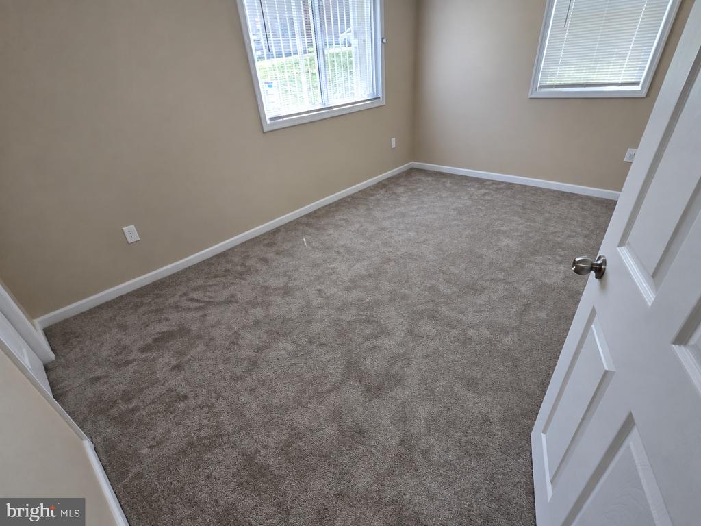 Vacant bedroom with gray-brown carpet, beige walls, and two windows with blinds.