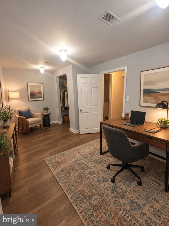 Staged home office with a wooden desk, laptop, and patterned rug; an open closet is visible behind it.